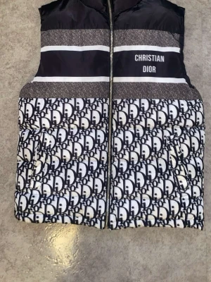 Christian Dior Väst  - Säljer en dunväst från Christian Dior med svartvitt logomönster och tydlig Christian Dior-text på bröstet. Västen har quiltad design och dragkedja framtill. Materialet är syntet och polyester, med en glansig finish på insidan. Perfekt för dig som gillar statement-plagg.