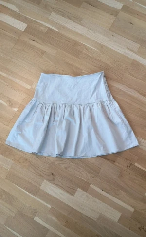 Ljusbeige kortkjol från Holly & Whyte - En ljusbeige kortkjol från Holly & Whyte med volang nedtill. Kjolen har en markerad midja och dragkedja i sidan för enkel på- och avtagning.