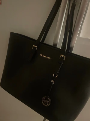 Svart axelväska från Michael Kors - Köpt för 2199 kr. Priset går att diskuteras.Säljer en stilren svart axelväska från Michael Kors med klassisk MK-logga i metall. Väskan har en enkel, rektangulär form och är tillverkad i skinn med guldfärgade detaljer. Perfekt för dig som gillar snygga och praktiska accessoarer.