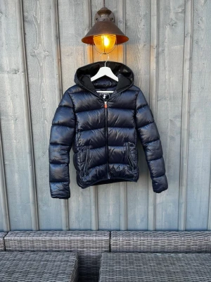 Save the duck Jacka - Snygg mörkblå pufferjacka med huva och dragkedja framtill. Jackan har två praktiska fickor med dragkedja och ett glansigt yttermaterial som ger en cool look. Perfekt för kalla dagar och enkel att matcha med dina favoritkläder. Strl: 14y