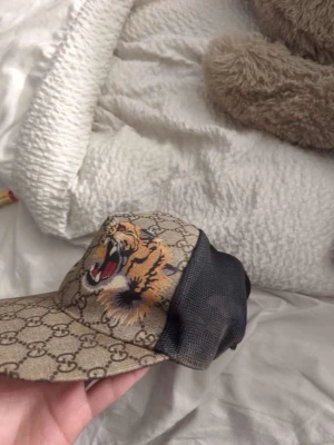 keps med tiger - Säljer en snygg keps med klassiskt GG-mönster i beige och brunt, broderad tiger framtill och svart mesh på sidorna. Justerbar rem med gröna och röda ränder samt guldfärgad ring. Perfekt för dig som vill sticka ut med lyxig streetstyle.
