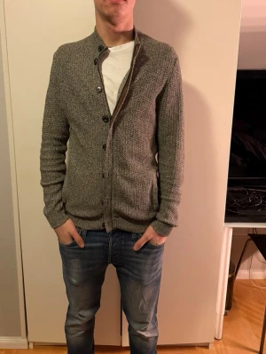 Grå kofta från Massimo Dutti, M - Snygg gråmelerad kofta från Massimo Dutti i bomull. Modellen har lång ärm, ribbade muddar och knappar framtill. Perfekt för dig som gillar stilrena och klassiska plagg med lite struktur. Passar dig som vill ha en normal passform och gillar neutrala färger.