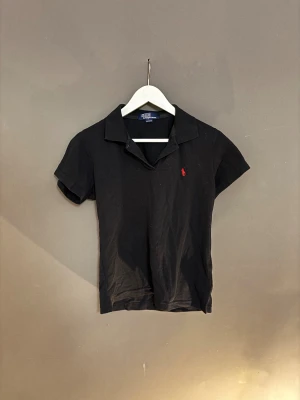 Svart pikétopp från Polo Ralph Lauren - Klassisk svart pikétopp från Polo Ralph Lauren med korta ärmar och krage. Toppen har en röd broderad logga på bröstet och är tillverkad i 100% bomull. Perfekt för en stilren och sportig look.