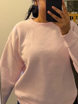Ljusrosa sweatshirt i bomull - Enkel och stilren ljusrosa sweatshirt med rund hals och ribbade muddar. Tröjan har en loose passform och är tillverkad i mjuk bomull, perfekt för chill dagar eller när du vill ha något bekvämt på dig.