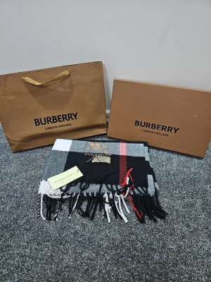 Rutig kashmirväst från Burberry - Säljer en snygg rutig väst från Burberry i 100% kashmir. Västen har klassiskt rutmönster i svart, grått, vitt och en röd detalj, samt broderad Burberry-logga framtill. Mjukt material och fransar nedtill ger en lyxig känsla.