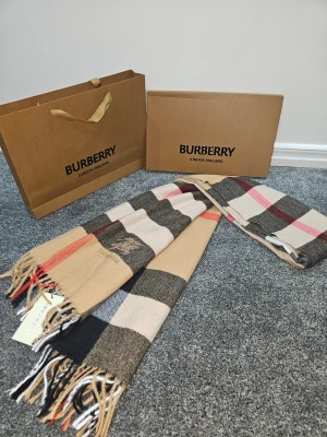 Rutig Burberry ullväst i beige och svart - Säljer en klassisk rutig väst från Burberry i mjuk ull. Västen har breda rutor i beige, svart, vitt och röda detaljer samt broderad Burberry-logga med riddare. Materialet är ull och designen har fransar i nederkant. Perfekt för dig som gillar ikoniska mönster och lyxig känsla.