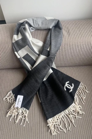 Grå och vit Chanele halsduk med fransar - Snygg halsduk från Chanele i grått och vitt med stora grafiska mönster och ikoniska C-loggan. Halsduken har långa vita fransar i ändarna och känns riktigt lyxig. Perfekt att slänga över axlarna för en cool look.