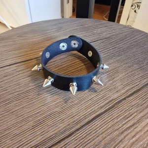 Svart punk nitad läderchoker med spikes - Srmband punk svart choker i läderimitation i PU med silvriga spikes och tryckknappar. 22 cm långt. Perfekt accessoar för dig som gillar edgy stil och vill sticka ut. Justerbar passform med två knappar. Snygg att matcha med streetwear eller rockiga outfits.