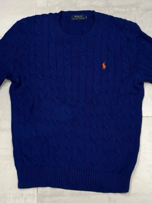 Blå stickad tröja från Polo Ralph Lauren - Snygg marinblå stickad tröja från Polo Ralph Lauren med klassiskt kabelstickat mönster och orange broderad logga på bröstet. Rund halsringning och ribbade muddar vid ärmslut och nederkant. Perfekt för dig som gillar stilrena och tidlösa plagg.