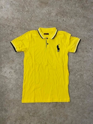 Gul piké från Polo Ralph Lauren - Säljer en gul pikétröja från Polo Ralph Lauren med svart broderad logga på bröstet. Tröjan har korta ärmar, krage med svarta och vita ränder samt knappar framtill. Materialet är mjuk bomull och passformen är normal.