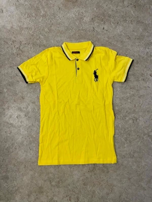 Gul piké från Polo - Säljer en gul pikétröja från Polo med klassisk krage och korta ärmar. Tröjan har svarta och vita ränder på krage och ärmslut samt en stor svart Polo-logga broderad på bröstet. Perfekt för dig som gillar sportig och färgstark stil.