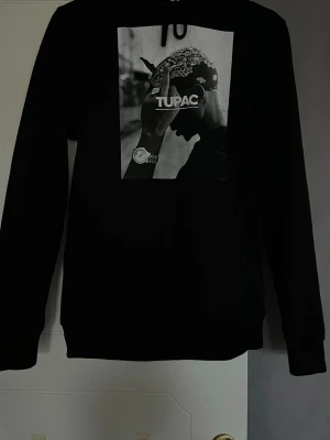 Svart Tupac t-shirt Mister Tee S - Svart t-shirt från Mister Tee x Artists i storlek S med stort tryck av Tupac på framsidan. T-shirten har klassisk passform och rund hals. Materialet är mjuk bomull och trycket är i svartvitt med tydlig Tupac-logga. Perfekt för dig som gillar streetwear och hiphop.