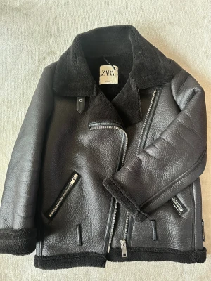 Svart pilot pälsjacka från Zara - Säljer en svart pilot pälsjacka från Zara med grov struktur och mjukt foder. Jackan har bred krage, flera dragkedjor och detaljer i skinnimitation. Perfekt för kalla dagar och ger en cool look med sina markerade sömmar och rejäla muddar.