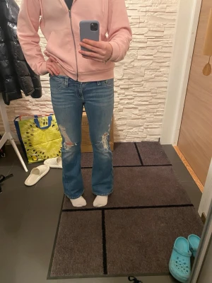 Blå bootcut jeans med slitningar - Säljer ett par blå bootcut jeans med snygga slitningar på framsidan av benen och coola fickdetaljer bak med knappar. Jeansen har låg midja och är gjorda i klassiskt jeansmaterial. Perfekt för dig som gillar en avslappnad och trendig look. Obs de har inga framfickor 