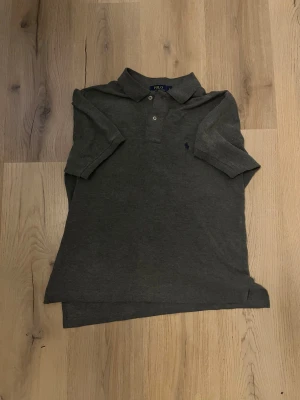 Mörkgrå pikétröja från Polo Ralph Lauren - Säljer en mörkgrå pikétröja från Polo Ralph Lauren i storlek L, custom fit. Tröjan har klassisk krage, korta ärmar och två knappar framtill. Materialet är mjukt och känns som bomull. Diskret logga broderad på bröstet.
