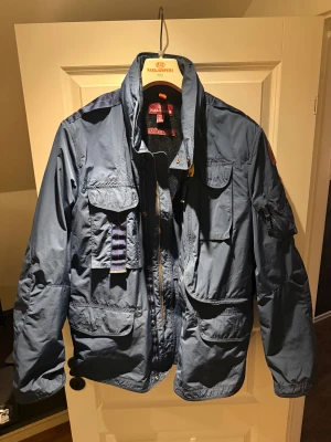 Blå vindjacka från Parajumpers XL - Snygg blå vindjacka från Parajumpers i storlek XL. Jackan har flera praktiska fickor, dragkedja och coola detaljer som gul rem och patch på ärmen. Materialet är lätt polyester och jackan har långärmad design med hög krage. Perfekt för dig som gillar street och funktion. Halvår gammal. Passar L-XL. 