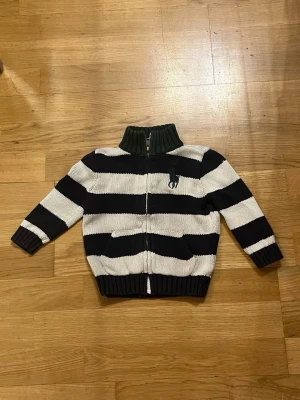 Randig kofta från Ralph Lauren - Säljer en snygg stickad kofta från Ralph Lauren med breda svarta och vita ränder för barn. Koftan har dragkedja framtill, ribbad krage och muddar samt två fickor. Perfekt för dig som gillar klassisk stil med en sportig touch.