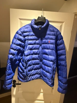 Blå pufferjacka från Polo Ralph Lauren - Säljer en blå pufferjacka från Polo Ralph Lauren med horisontella quiltade sektioner och dragkedja framtill. Jackan har ståkrage, två sidofickor med dragkedja och den klassiska röda Polo-loggan på bröstet. Perfekt för kyliga dagar.