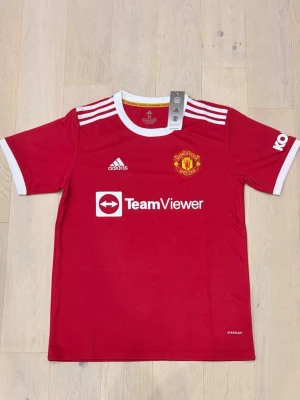 Manchester United adidas matchtröja - Röd Manchester United matchtröja från adidas med vita detaljer och klubbmärke på bröstet. Tröjan har TeamViewer-tryck på framsidan, vita ränder på axlarna och vit krage. Tillverkad i polyester med Aeroready-teknologi för extra komfort.