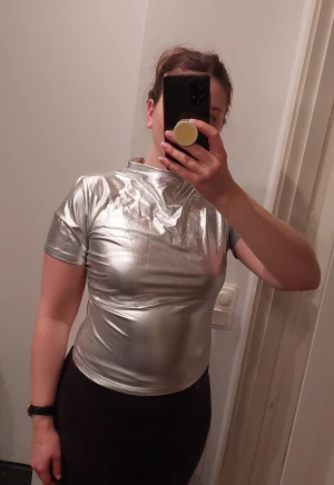 Silvrig topp - Silvrig topp med halterneck och veckad söm över ena axeln och ena sidan. Elastisk och festlig! Stl. M men passar även S. Behöver strykas något, annars inga anmärkningar.