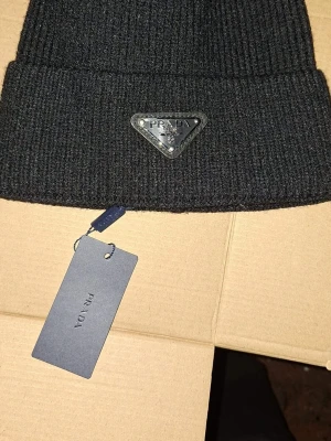 Svart stickad mössa från Prada - Snygg svart mössa från Prada med ribbad stickning och metallmärke framtill. Mössan har en klassisk uppvikt kant och är tillverkad i mjukt material som håller dig varm. Perfekt accessoar för att lyfta din look med en lyxig touch.