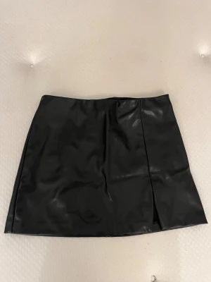 Svart kort kjol i skinn från DIVIDED - Säljer en svart kort kjol från DIVIDED i skinnimitation. Kjolen har en stilren design med en liten slits framtill och sitter snyggt i midjan. Perfekt för dig som gillar en edgy och trendig look.