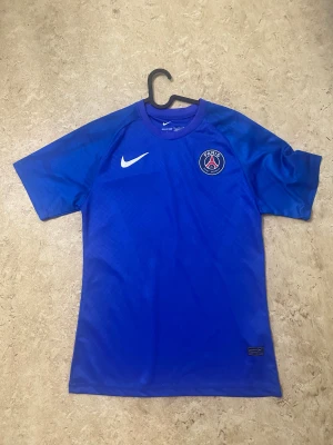 Blå PSG x Nike fotbollströja - Säljer en blå Paris Saint-Germain fotbollströja från Nike med korta ärmar och rund hals. Tröjan har PSG-logga på bröstet och Nike Swoosh i vitt. Materialet är lätt polyester med subtilt rutmönster. Perfekt för dig som älskar fotboll och vill ha en snygg supportertröja.