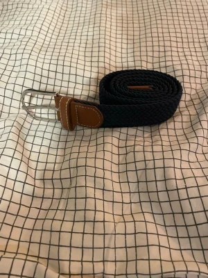 Mörkblå flätad bälte med skinn - Snyggt mörkblått flätat bälte med detaljer i brunt skinn och vit söm. Spännet är i silverfärgad metall och bältet har en flexibel, vävd struktur som gör det bekvämt att bära. Perfekt för att ge din outfit en stilren touch.