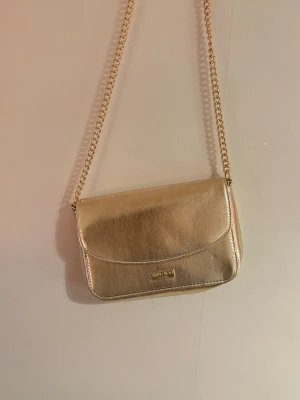 Guldig axelväska från Jimmy Choo - Säljer en snygg guldig axelväska från Jimmy Choo Parfums med kedjerem. Väskan har en stilren, rektangulär form och är tillverkad i skinnliknande material med metallisk finish. Perfekt för dig som vill ha en statement piece med lyxig vibe.