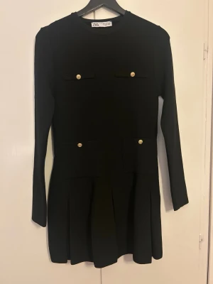 Svart kort klänning från Zara - Supersnygg svart kort klänning från Zara med lång ärm och rund hals. Klänningen har guldfärgade knappar på fickorna framtill. Endast använd 1 gång. 