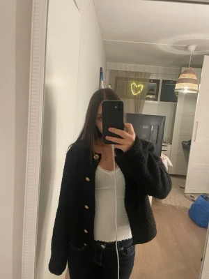 Svart kofta med guldknappar - Mysig svart kofta med guldknappar framtill. Koftan har en lite fluffig och stickad känsla, perfekt att slänga på sig när det är kyligt. Långärmad och med en klassisk rund halsringning. Passar till jeans eller kjol för en chill look.