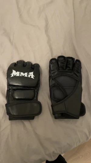  MMA handskar - Säljer ett par svarta MMA handskar med öppen hand och fingrar. Handskarna har vit MMA-logga på ovansidan och bred kardborreknäppning runt handleden. Tillverkade i syntetmaterial med vaddering för extra skydd och komfort under träning. Dom är som ny.
