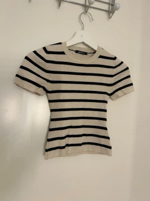Randig topp från Gina Tricot XS - Säljer en beige och svart randig topp från Gina Tricot i storlek XS. Toppen har rund halsringning och korta ärmar. Materialet är stickat och ger en mjuk känsla. Passformen är slim och toppen har ett klassiskt, tidlöst utseende.