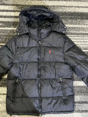 Svart pufferjacka från Polo Ralph Lauren - Svart pufferjacka från Polo Ralph Lauren med huva och flera dragkedjeförsedda fickor. Jackan har ett rött Polo-logo broderat på bröstet och är tillverkad i syntetmaterial. Perfekt för kalla dagar och har en schysst, lite oversized passform.