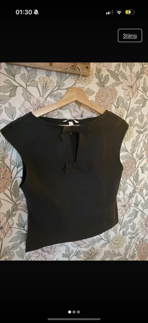 Svart ärmlös topp med rosetter - Säljer en svart, stilren topp med breda axlar och två dekorativa rosetter framtill. Toppen är ärmlös och har en rak, något boxig passform. Materialet känns slätt och har en lätt glans. Perfekt för dig som gillar minimalistisk design med en söt detalj.