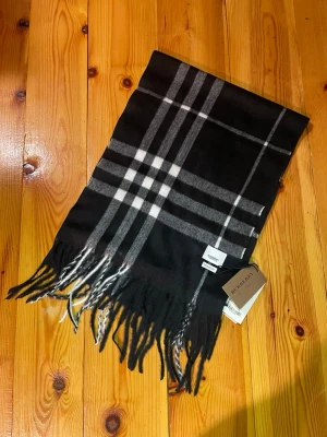 Svart rutig Burberry scarf i kashmir - Snygg svart halsduk från Burberry med klassiskt rutigt mönster i vitt och grått. Scarfen är tillverkad i 100% kashmir och har fransar längs nederkanten. Perfekt för dig som vill ha en lyxig och tidlös look med premiumkänsla.