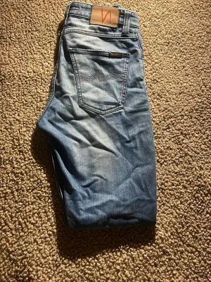 Ljusblå slim jeans från Nudie Jeans - Säljer ett par ljusblå slim jeans från Nudie Jeans med klassisk femficksdesign och snygga slitningar. Jeansen har en smal passform och är tillverkade i mjukt bomullsmaterial med Nudie-logga på läderpatchen bak. Perfekt för dig som gillar en clean och modern look.