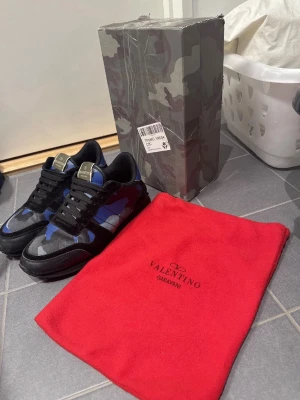 Valentino Garavani Rockrunner sneakers - Säljer ett par Valentino Garavani Rockrunner sneakers med blått och svart camouflage-mönster. Skorna har rund tå, snörning och platt sula. Kommer med originalkartong i camouflage och röd dustbag med Valentino-logga. Säljs då de inte kommer till stor användning, kom gärna med byten!