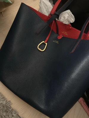 Mörkblå och röd shoppingväska Lauren - Säljer en stor och rymlig shoppingväska från Lauren Ralph Lauren i mörkblått och rött skinn. Väskan har guldfärgad detalj framtill och röda handtag. Perfekt för dig som vill ha plats för allt och samtidigt se snygg ut.