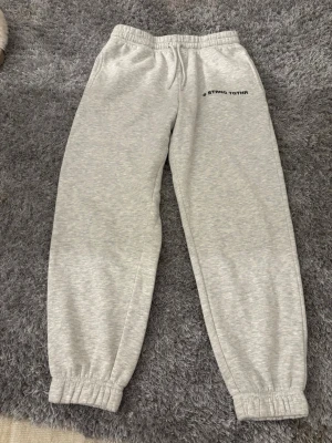 Grå mjukisbyxor från Newyorker  - Grå mjukisbyxor från FB Sister i storlek S med trycket '@STRNG TGTHR' på ena benet. Byxorna har resår och snörning i midjan samt muddar vid bensluten. Tillverkade i mjukt bomullsmaterial som är perfekt för chill och häng. 