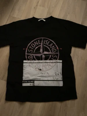 Svart Stone Island t-shirt med tryck - Säljer en svart t-shirt från Stone Island i storlek M. T-shirten har ett stort, lila tryck med logga på bröstet och en vit grafisk detalj med text och slitna effekter. Klassisk rund hals och korta ärmar. Perfekt för dig som gillar streetwear och vill sticka ut.