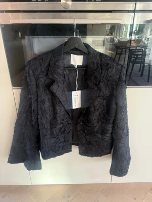 Svart strukturerad blazer EU 40 helt ny! - Säljer en helt ny svart, åtsittande blazer med strukturerat mönster och cool textur. Jackan har klassisk krage, långa ärmar och knappar framtill. Perfekt för dig som vill ha en unik och trendig look med extra edge.