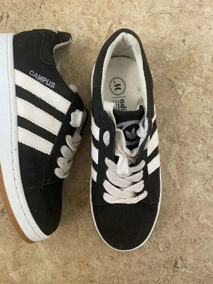 Adidas Campus svarta sneakers - Svarta Adidas Campus sneakers med klassiska vita ränder på sidorna och vit sula. Skorna har rund tå, platt sula och snörning med vita skosnören. Ovandelen är i mocka och insidan är fodrad i textil. Perfekt för dig som gillar retrostil och streetwear.