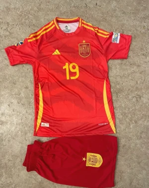 Spaniens fotbollströja och shorts - Säljer ett rött fotbollskit från Adidas med Spaniens landslagströja och matchande shorts. Tröjan har gula detaljer, nummer 19 och namntryck Lamine Yamal på ryggen. Materialet är lätt polyester och har officiella landslagsemblem både på tröjan och shortsen.