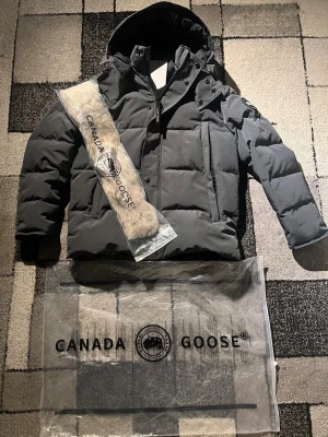 Canada Goose Wyndham  - Säljer en mörkgrå dunjacka från Canada Goose med huva och avtagbar pälskant. Jackan har dragkedja, tryckknappar och flera fickor framtill. Perfekt för kalla dagar och riktigt snygg streetstil.