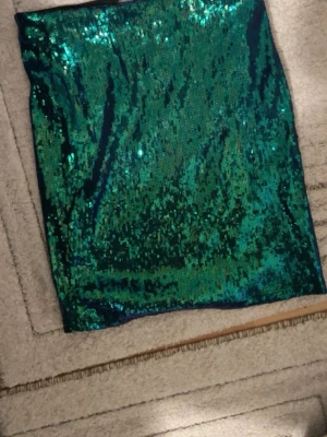 Grön glittrig paljettkjol - Säljer en kort kjol täckt med gröna paljetter som ger ett riktigt glittrigt och festligt intryck. Kjolen har en rak och tight passform som sitter snyggt på kroppen. Perfekt för dig som vill sticka ut med lite extra bling och färg.