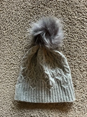 Grå stickad mössa med pälsboll - Säljer en snygg grå stickad mössa med ett mönstrat strukturstickat parti och en stor fluffig pälsboll på toppen. Mössan har bred ribbad kant och är perfekt för kalla dagar. Passar dig som vill ha både stil och värme.