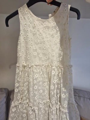 Vit blommig ärmlös barnklänning - Superfin vit barnklänning med blommigt mönster och ärmlös design. Klänningen har volangdetaljer och ett lager av transparent tyg med broderade blommor. Perfekt för sommarens alla tillfällen.