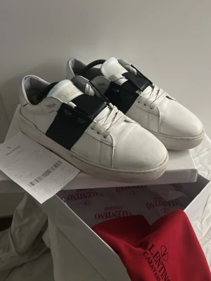 Valentino Garavani Open sneakers vit/svart - Säljer ett par Valentino Garavani Open sneakers i vitt skinn med bred svart läderdetalj över sidan. Klassisk rund tå, platt sula och snörning framtill. Kommer med originalkartong, dustbag och äkthetsbevis. Perfekt för dig som gillar lyxiga och stilrena sneakers.