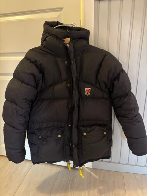 Svart dunjacka från Fjällräven - Svart dunjacka från Fjällräven med hög krage och huva. Jackan har gul snörning nedtill, två stora fickor med knappar och dragkedja framtill. Klassisk Fjällräven-logga på bröstet och puffig design som håller dig varm under kalla dagar. Storleken är stor i storlek, jag bär s och är 158 och den är lite för stor. Använd men i gott skick.